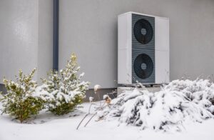 an outdoor heat pump unit surrounded by snowy grass and bushes
