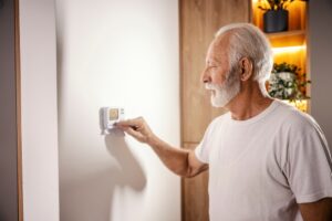 a senior man with gray hair and beard is adjusting the thermostat on the wall to turn his home's heat off